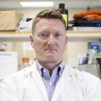 Dr Blair Leavitt has short light coloured hair. He is wearing a white lab coat.